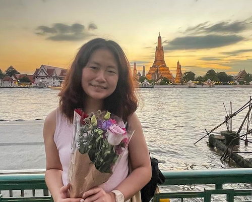 A smiling exchange student holding a bouquet of flowers poses in front of the iconic Wat Arun at sunset during her exchangesemester at CSII in Bangkok.